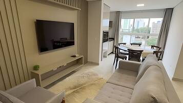 A modern living room with a flat-screen TV, a sofa, a dining area with a table and chairs, and a view of the city through large windows.