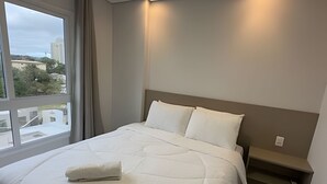 A hotel room with a large bed, a nightstand, a window with curtains, and a view of buildings and trees outside. - Apartamento no centro de Foz do Iguaçu (Foz do Iguaçu)