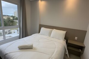 Una habitación de hotel con una cama grande, una mesita de noche, una ventana con cortinas y vistas a edificios y árboles.