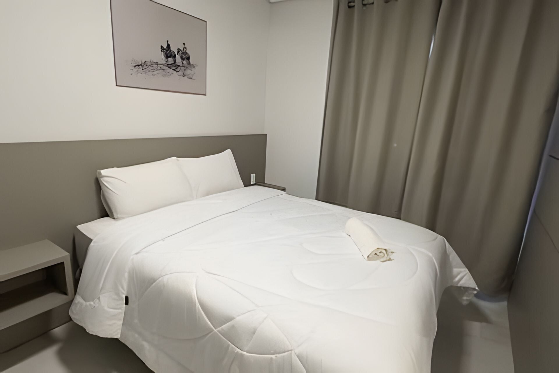 Et hotelværelse med seng, et natbord, et billed der sidder på væggen, samt et vindue med gardiner.