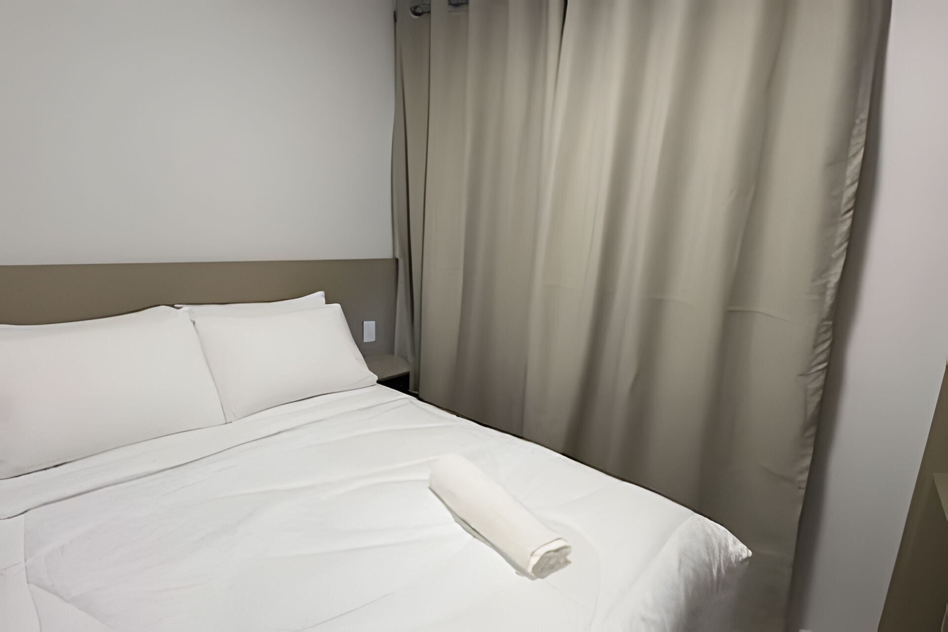 A neatly made bed with white linens and a rolled towel, a headboard, and a curtain.