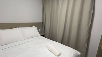 A neatly made bed with white linens and a rolled towel, a headboard, and a curtain.