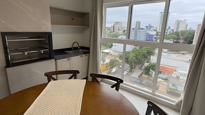 A room with a dining table, chairs, a kitchen area, and a large window with a city view. - Apartamento no centro de Foz do Iguaçu (Foz do Iguaçu)