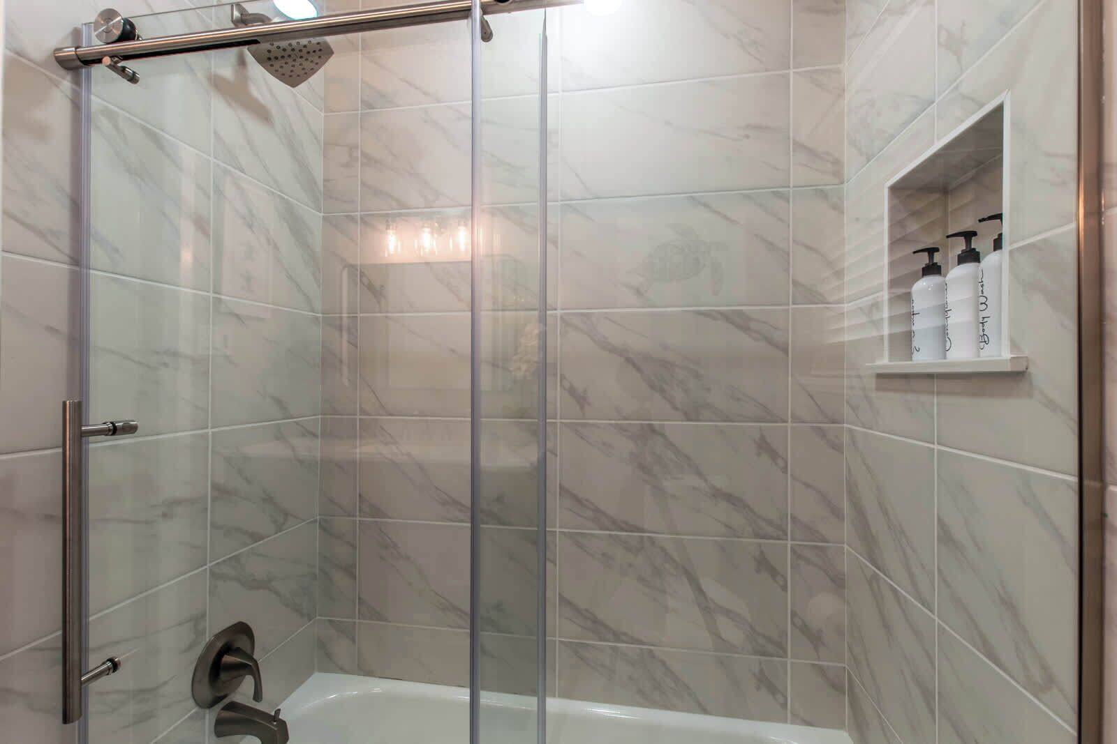 A modern bathroom with a glass-enclosed shower, a white bathtub, and a shelf with toiletries.