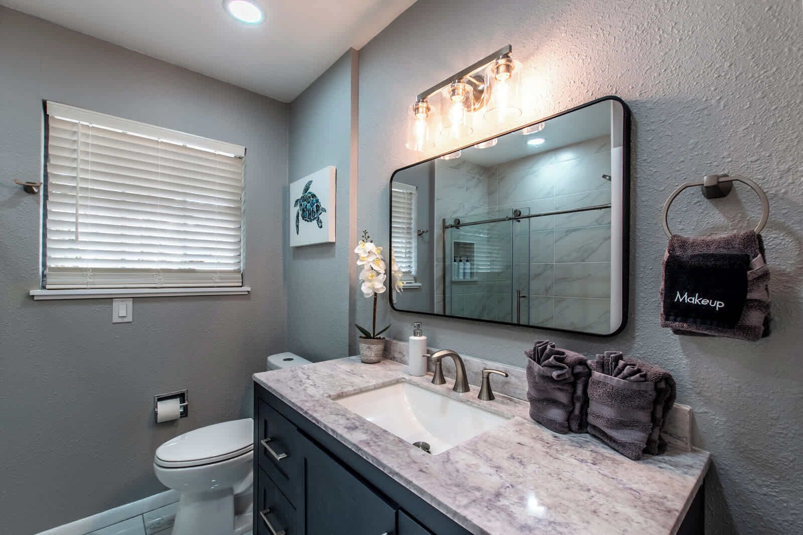 Une salle de bain dotée d’un grand miroir, d’un lavabo avec un plan de toilette en marbre, de toilettes et d’un porte-serviettes sur lequel est posée une serviette étiquetée « Maquillage ».