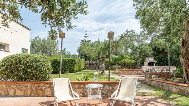 Un patio con dos sillas blancas y una mesa pequeña, rodeado de muros de piedra y vegetación.