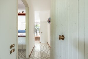 Un pasillo estrecho con una puerta blanca, suelo de baldosas y vista a una habitación con ventana y una mesa.