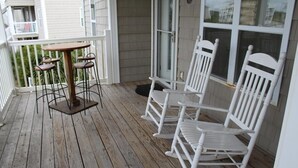 Una terraza de madera con una mesa y sillas, una baranda blanca y una ventana con vistas al exterior.