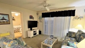 Una sala de estar con un sofá, una televisión, un ventilador de techo y una ventana con cortinas.