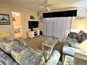 A living room with a sofa set, a television, a ceiling fan, and a window with curtains. - 3 Pools ! Condo at Ocean Isle Beach by Williamson Realty (Ocean Isle Beach)