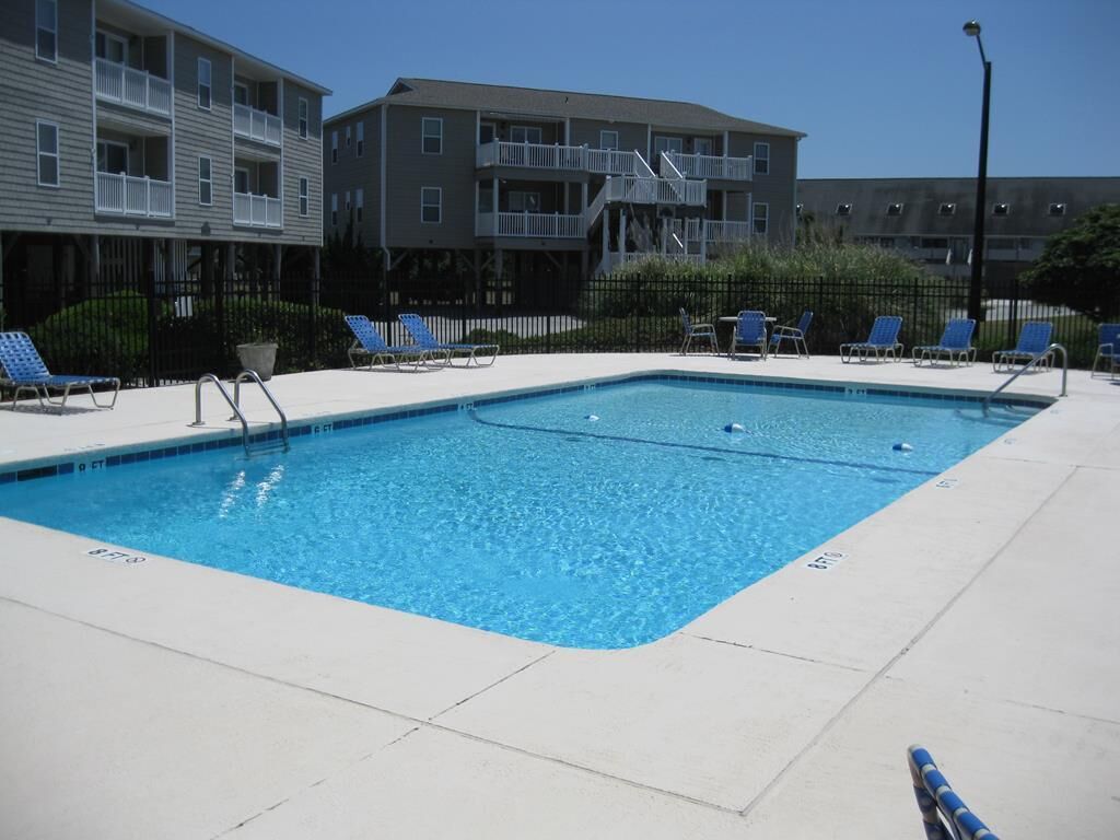 Une piscine rectangulaire à l’eau bleue, entourée d’une terrasse blanche. On y trouve des chaises longues et une petite table avec des chaises.
