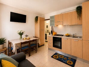 Eine kompakte Küche mit Holzausstattung, einem Esstisch mit Stühlen, einem an der Wand befestigten Fernseher und einem kleinen Blumentopf.