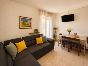 Ein Wohnzimmer mit einer Couch, einem Esstisch mit Stühlen, einem an der Wand befestigten Fernseher und einem Fenster mit Vorhängen.