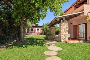 Una casa con un porche de piedra, un árbol y un edificio rojo al fondo.
