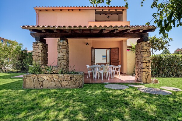 Un edificio de dos plantas con estructura de columnas de piedra, un patio con mobiliario exterior y un cielo despejado.