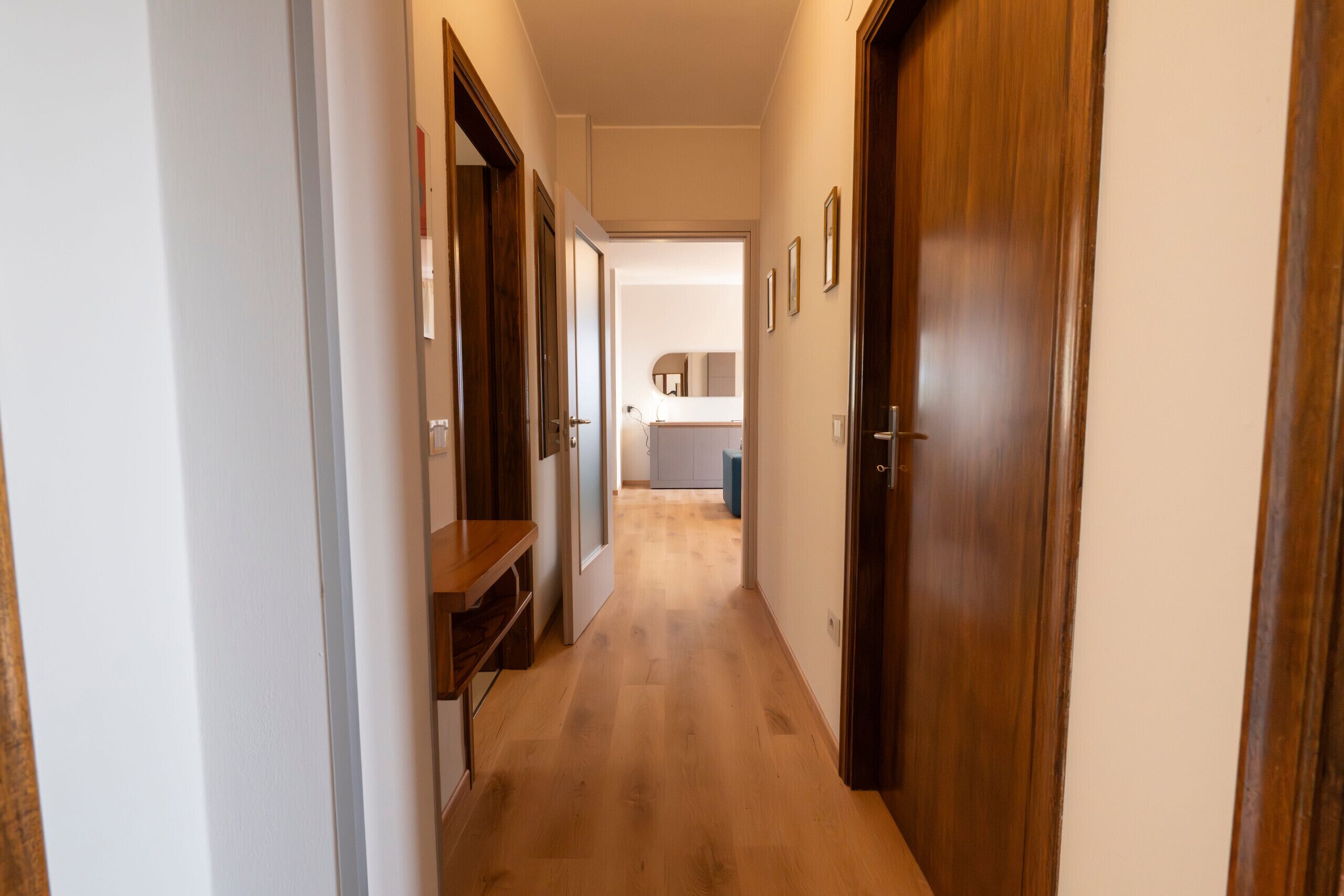 A hallway with wooden doors, a wooden bench, and a view into a room with a mirror and a sink.