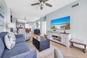 Ein Wohnzimmer mit einer Couch, einem Couchtisch und einem Fernseher. Im Hintergrund ist eine Küche zu sehen.