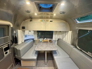 Een compact kampeerwagen interieur met een centrale tafel, beklede zitplaatsen, een kleine kitchenette en een raam.