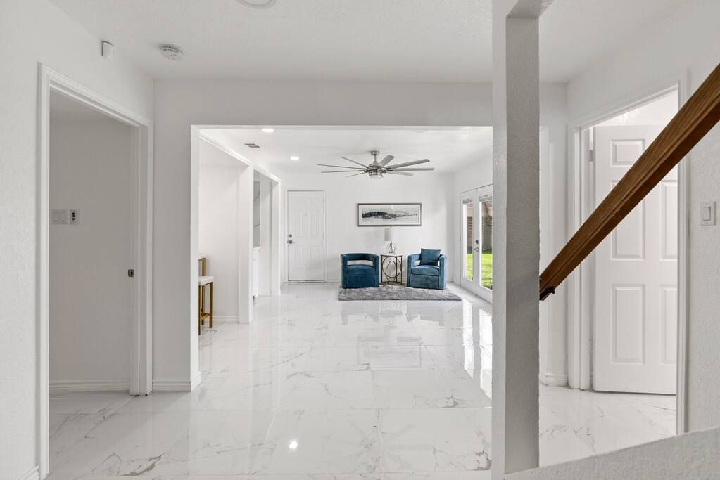A hallway with a marble floor, white walls, and a ceiling fan. There are two chairs with cushions and a small table with a vase.