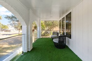 Eine überdachte Terrasse mit Kunstgrashalm, einem Korbstuhl-Garnitur und Blick auf die Straße und Bäume.