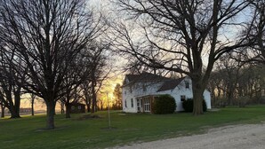 Une maison blanche avec un porche, entourée d’arbres et bordée d’une allée en gravier.