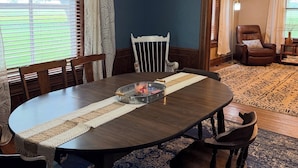 Une salle à manger avec une table en bois foncé, des chaises blanches, un lustre et une vue sur l’extérieur.