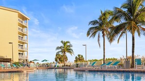 Ein Schwimmbecken mit Liegestühlen, Palmen und einem mehrstöckigen Gebäude.