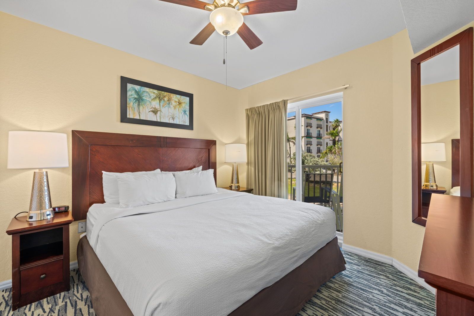 A hotel room with a bed, a ceiling fan, a nightstand with a lamp, a painting, and a view of buildings through a window.