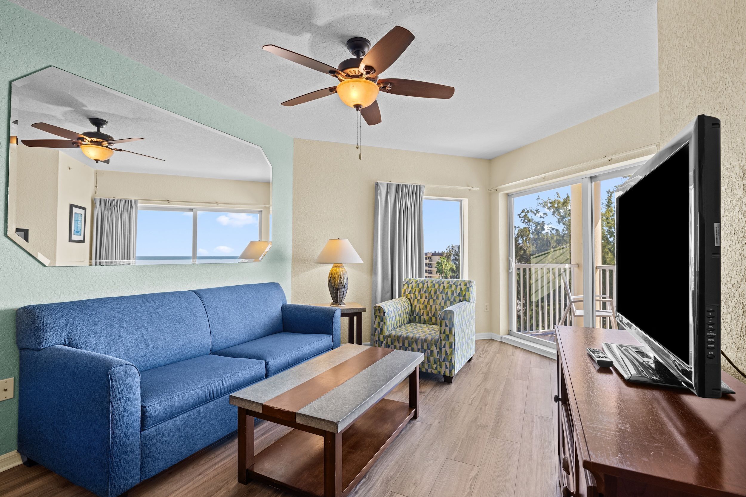 Ein Zimmer mit einem blauen Sofa, einem Holztisch, einem gemusterten Sessel, einem Deckenventilator, einem Fernseher und einem großen Fenster mit Blick.