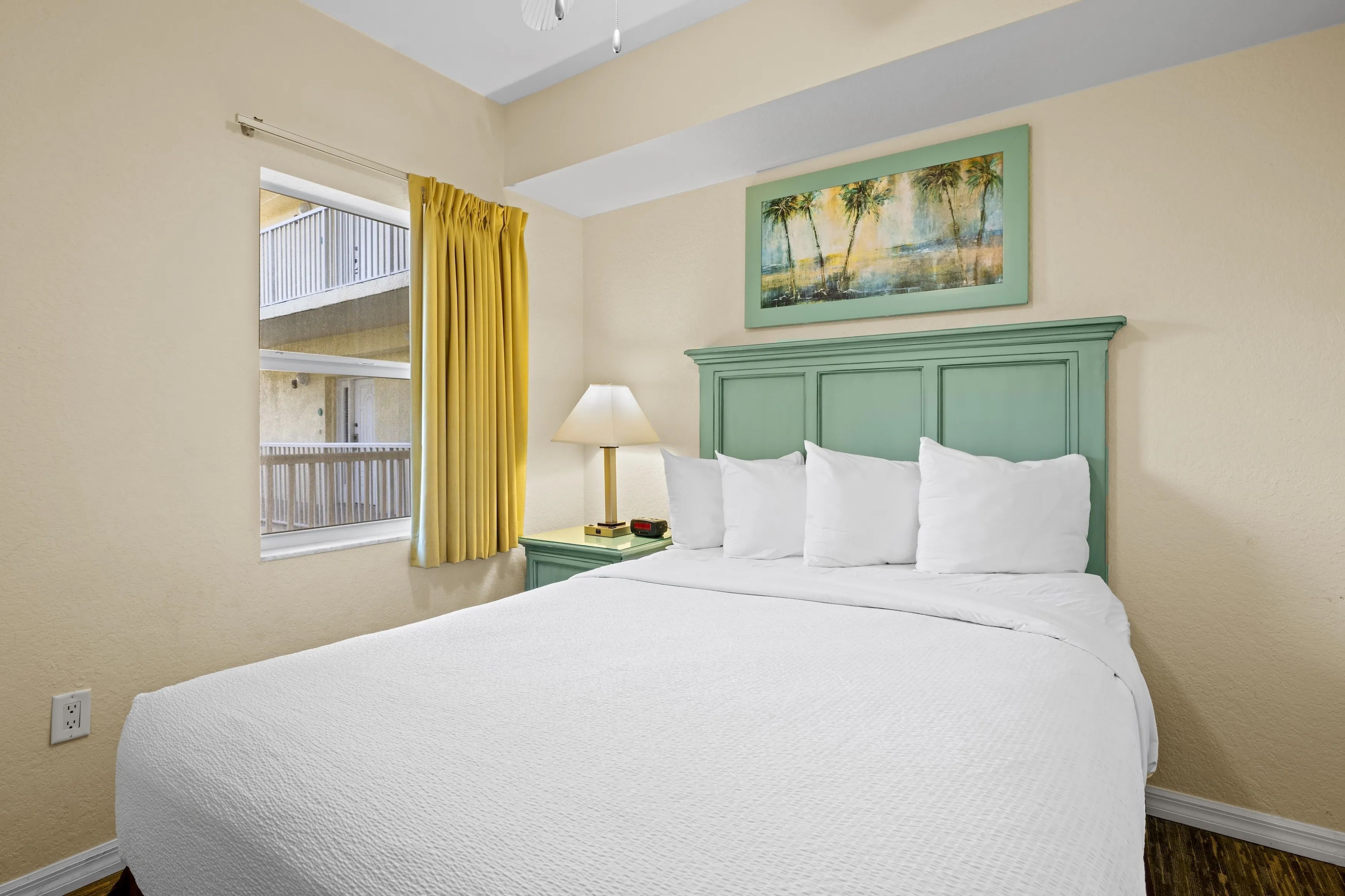 A hotel room with a bed, a nightstand, a lamp, a painting, and a window with yellow curtains.