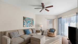 A living room with a sectional sofa, a round armchair, a ceiling fan, a TV, and a window with sheer curtains. - ADA Suite w/ Balcony & Beach Access (Treasure Island)