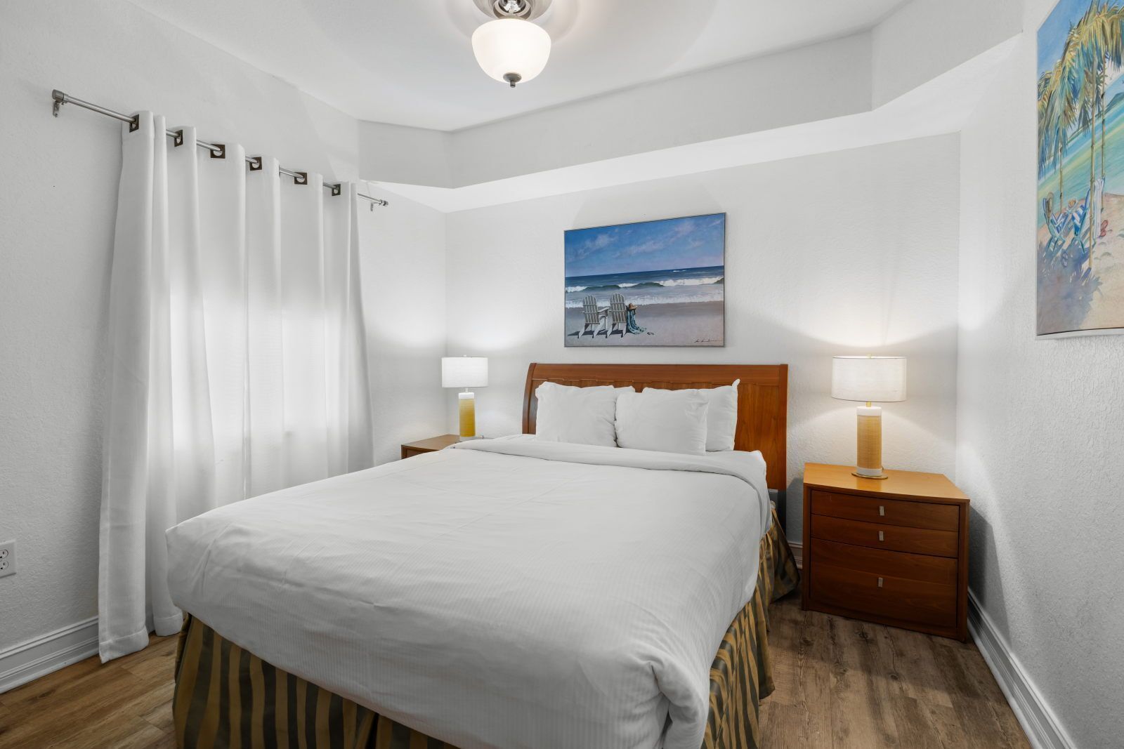 A hotel room with a large bed, two bedside tables with lamps, a painting of a beach scene, and a wooden floor.