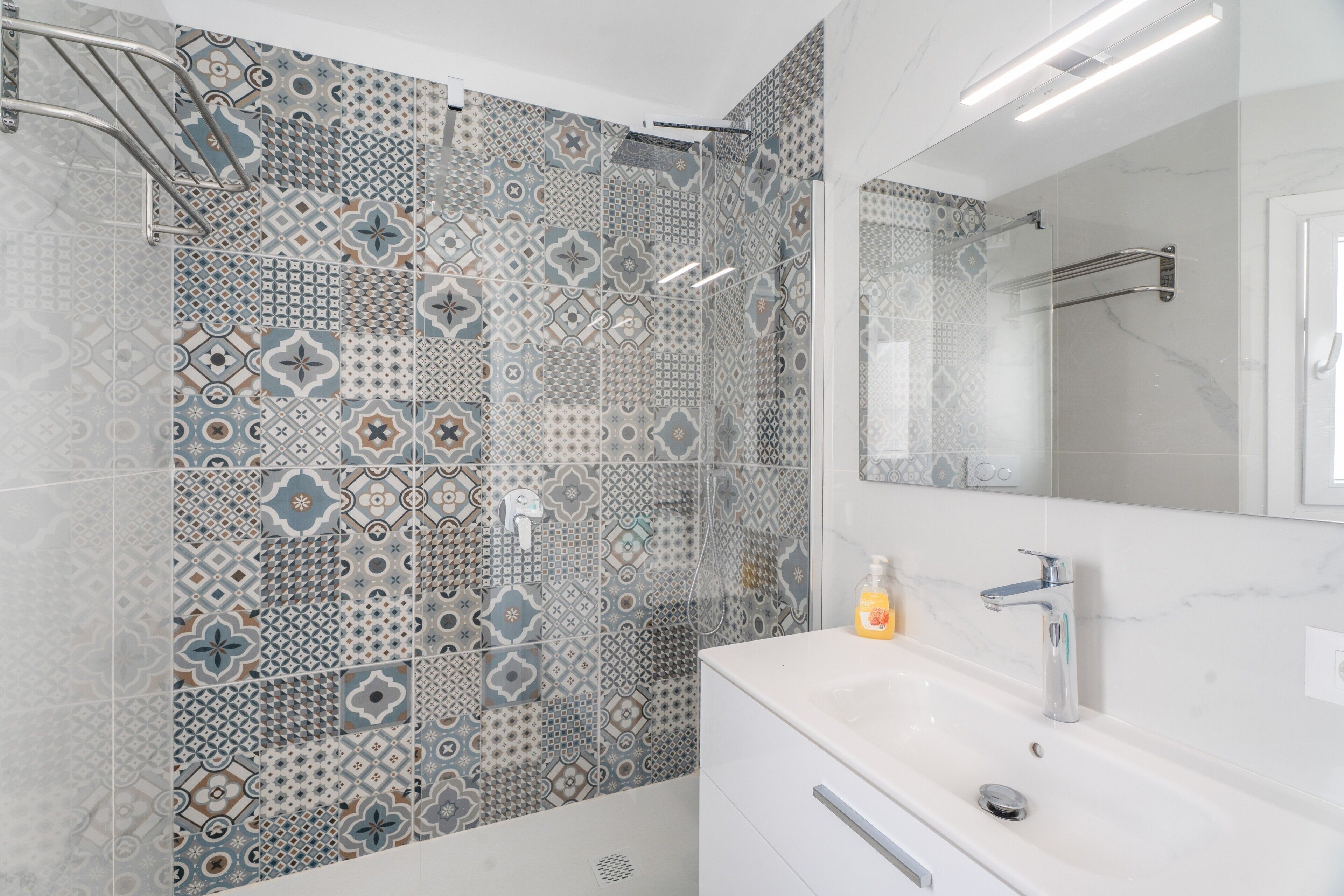 Une salle de bain avec des carreaux décorés, un lavabo blanc, un miroir et une douche.