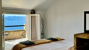 Een slaapkamer met een bed, uitzicht op zee, een balkon met een stoel en een vaas met takken.
