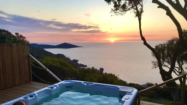 Een hottub op een houten terras met uitzicht op een wateroppervlak, met een zonsondergang op de achtergrond.