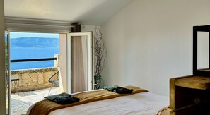 A bedroom with a bed, a view of the sea, a balcony with a chair, and a vase with branches.