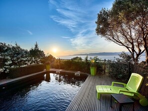 A wooden deck with a pool, a green chair, and a view of the sunset over the water.