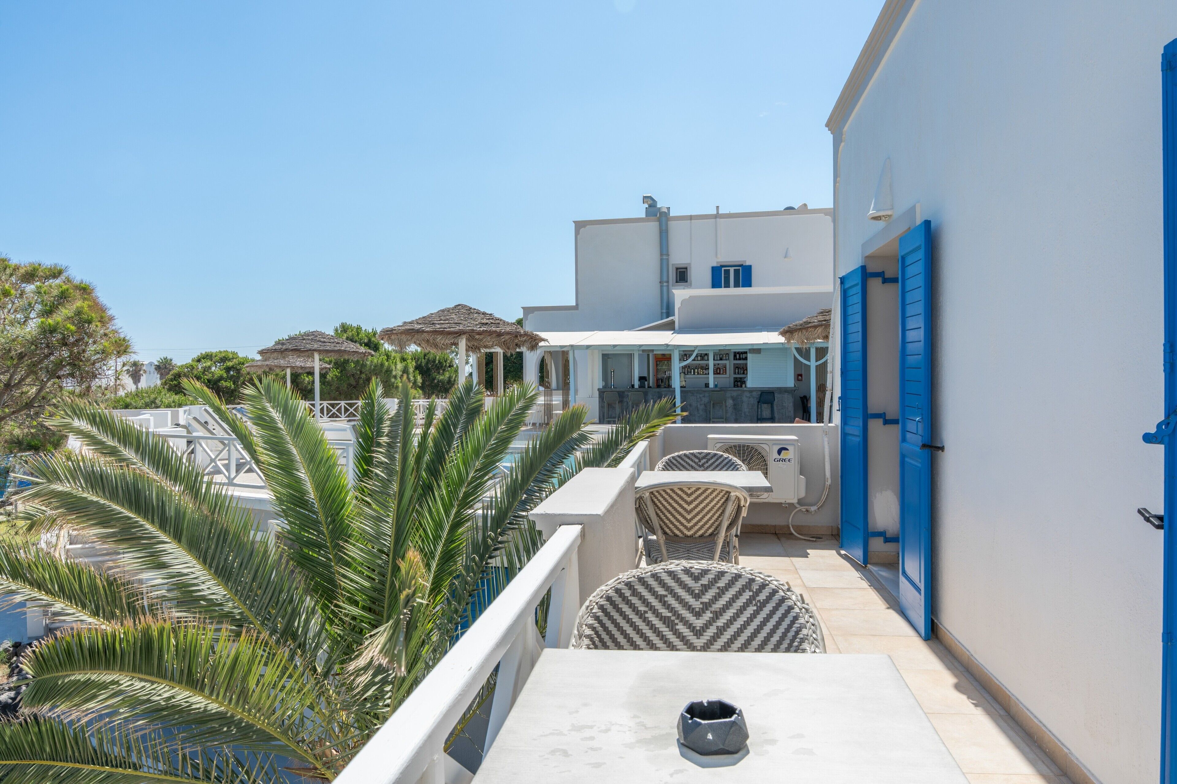 A balcony with a table, chairs, and a hat; a building with blue shutters; and a palm tree.