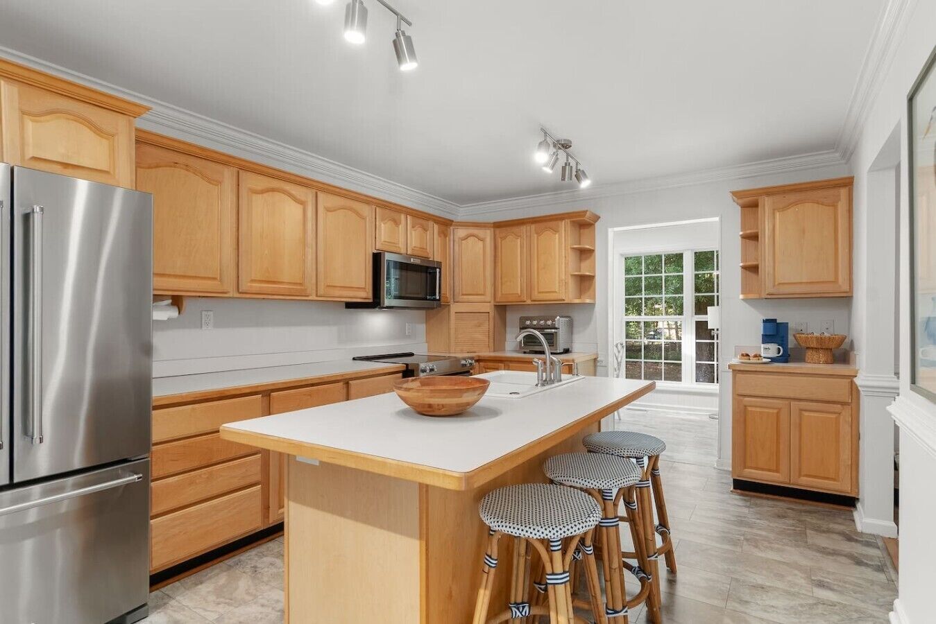 A modern kitchen with wooden cabinets, stainless steel appliances, a large island with stools, and a view to the outside.