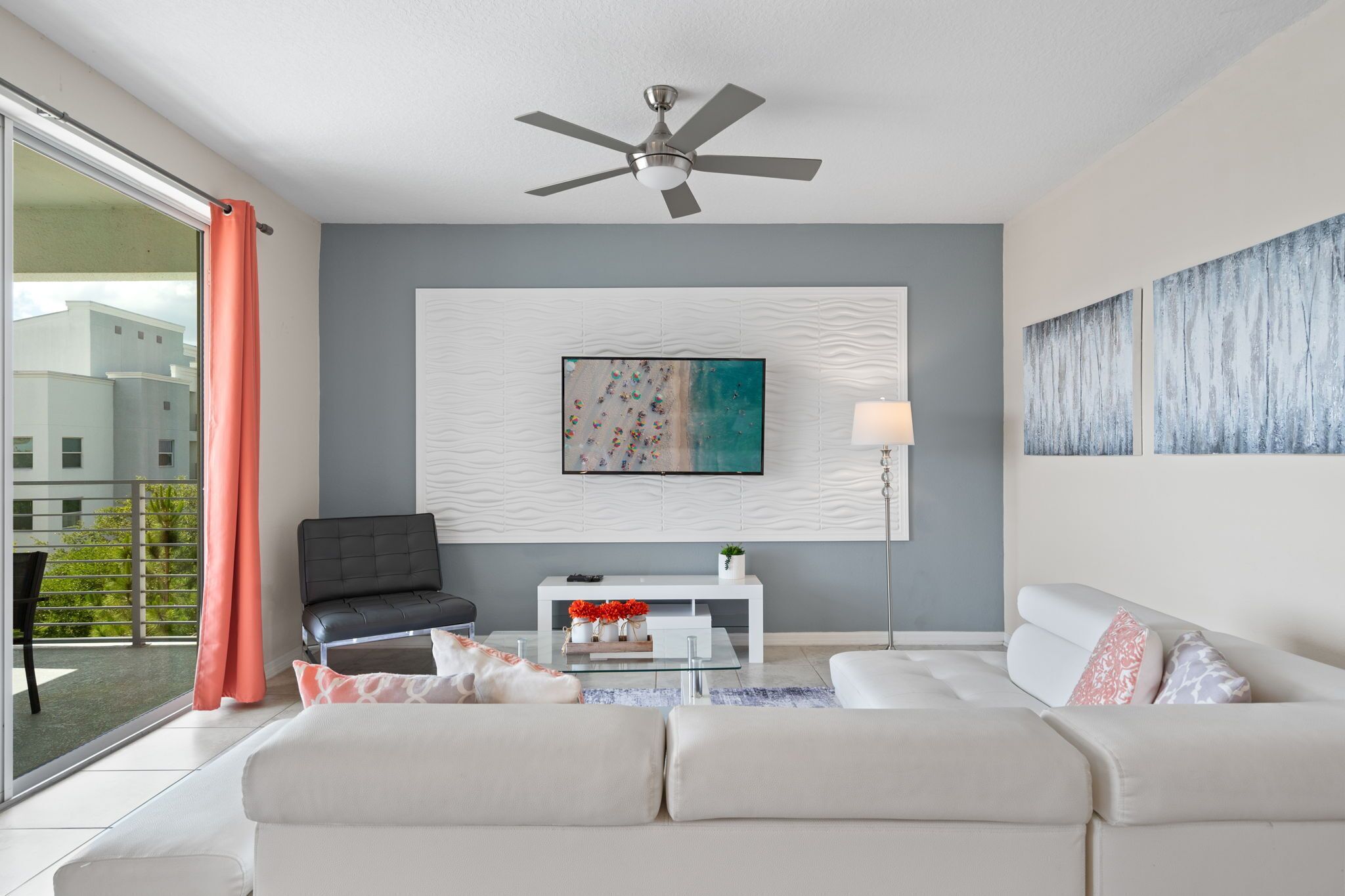Un salon moderne doté d’un ventilateur de plafond, d’un grand tableau abstrait, d’un canapé blanc, d’un fauteuil noir et d’une porte vitrée donnant sur un balcon.