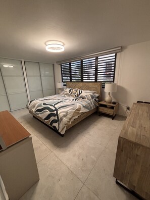 A bedroom with a bed, bedside tables, a desk, and a window with blinds. - Beach Front - Steps to beach 2-bdr remodeled Apt in Río Grande (Río Grande)