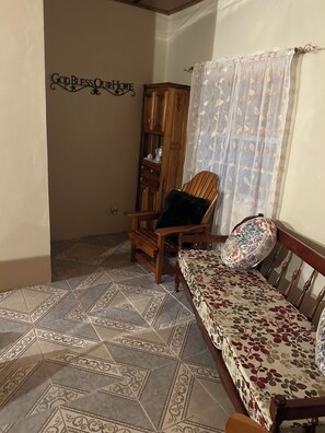 Ein Zimmer mit einem gemusterten Boden, einem Holzstuhl mit Kissen, einer Couch mit floralem Bezug, einem Holzkasten und einer Wand mit einer dekorativen Beschilderung.