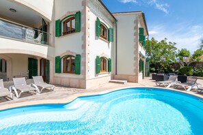 Un área junto a la piscina con sillones de descanso, una piscina y un edificio con contraventanas verdes.