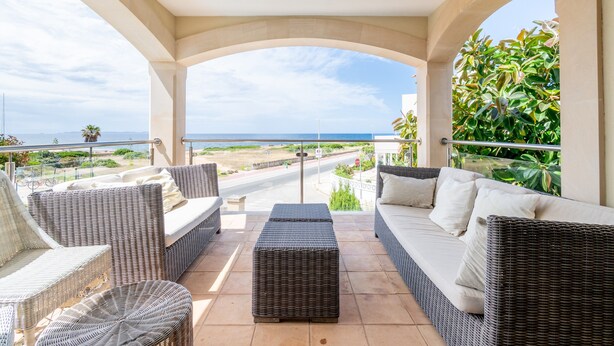 A covered outdoor seating area with wicker furniture, a view of the sea, and a paved pathway. - Mallorca Finca with Pool Directly on the Sea Es Trenc (Sa Ràpita)