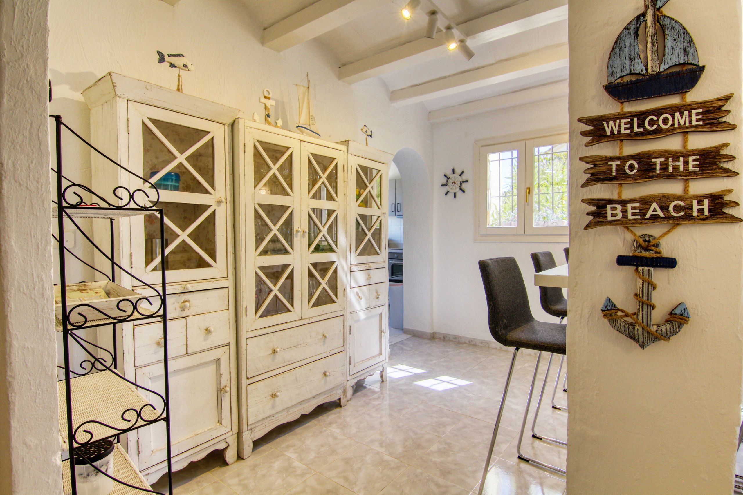 A room with a white cabinet with glass doors, a metal shelf, a wooden sign reading 'WELCOME TO THE BEACH', and a view of a beach through a window.