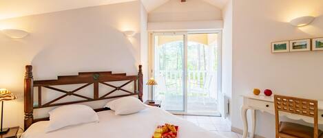 A bedroom with a large bed, a wooden headboard, a bedside table with a lamp, a chair, and a balcony with a view of greenery.
