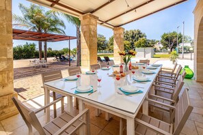 Ein überdachter Essbereich im Freien mit einem Tisch für sechs Personen, umgeben von Stühlen, mit Blick auf einen Pool und Palmen im Hintergrund.