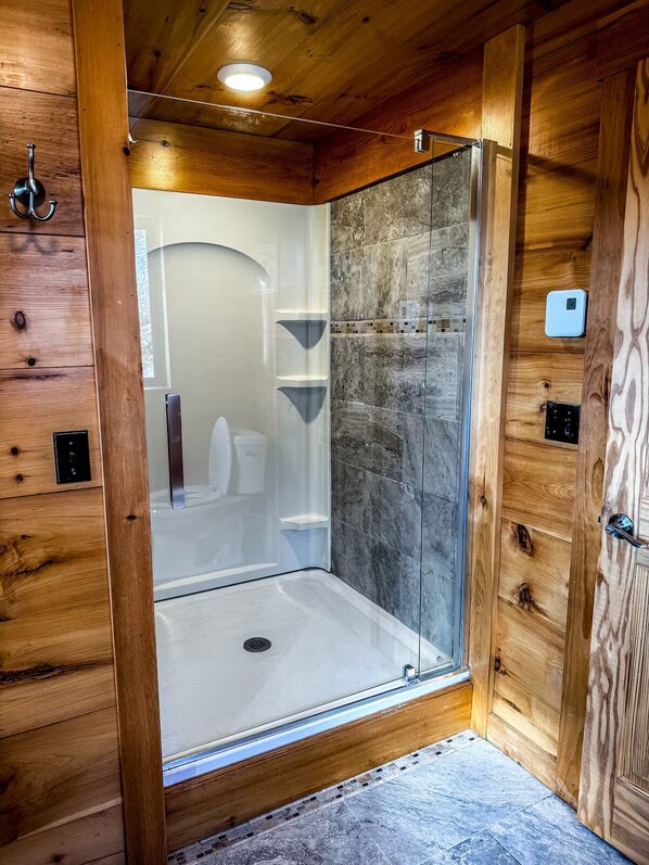 A wooden-paneled room with a glass-enclosed shower, a white toilet, and a small shelf.