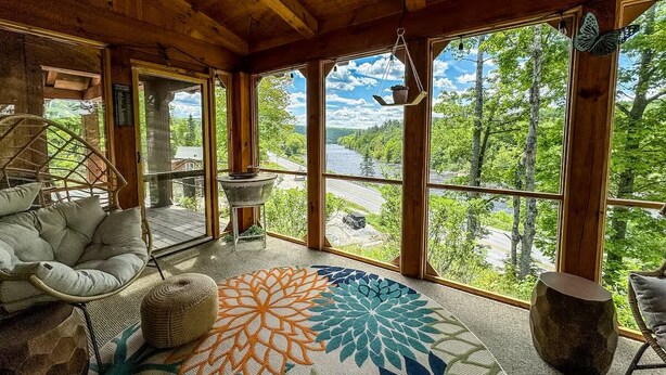 Ein gemütliches Zimmer mit großem Fenster, einem gewebten Sessel, einem runden Tisch und einem gemusterten Teppich.