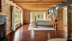 Ein Blockhaus-Interieur mit Kamin, Holzbalken, einem Bett und großen Fenstern.